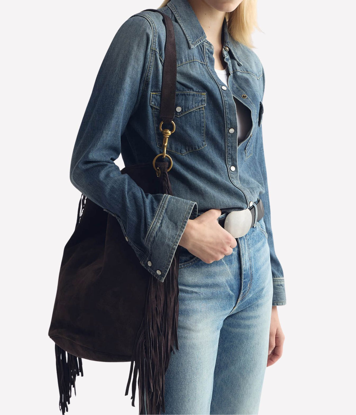 A model wearing a blue denim shirt and jeans, carrying a dark brown suede fringe bucket bag with gold-tone hardware over her shoulder, against a light gray background.