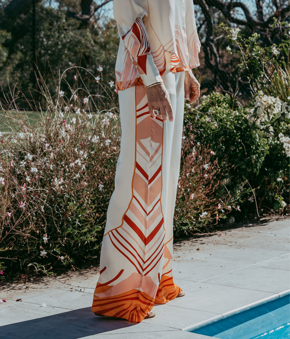 A person wearing a flowing cream shirt with orange and peach geometric patterns, standing on a tiled deck next to a swimming pool with garden foliage in the background.