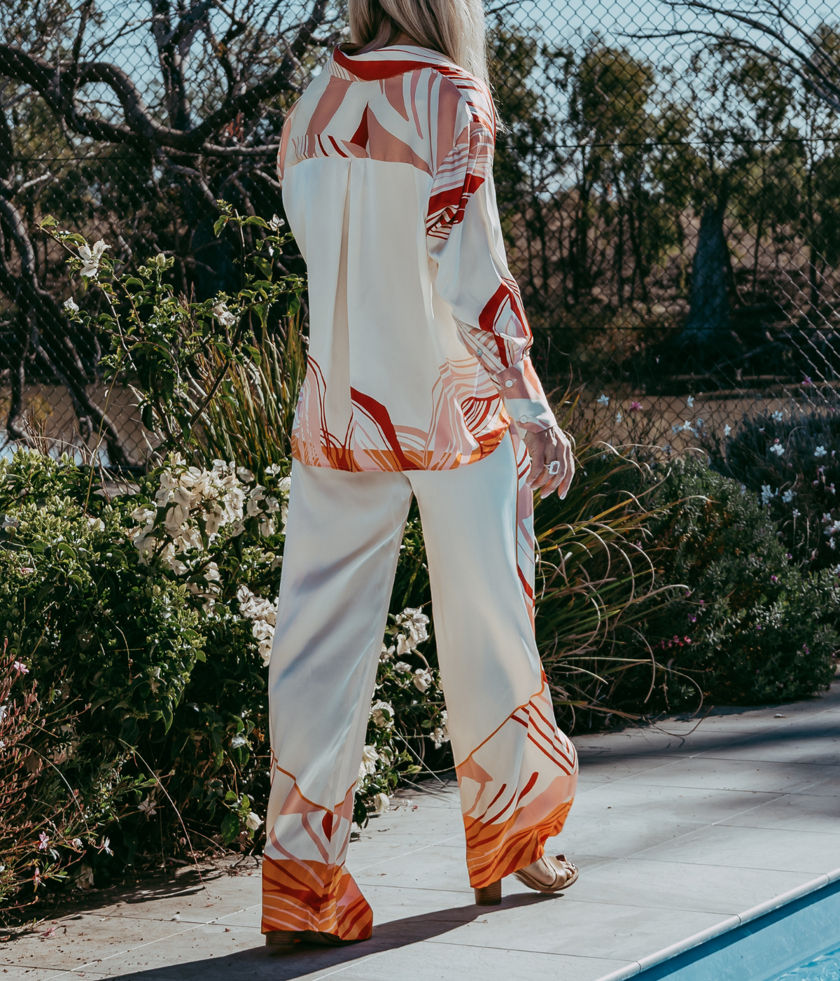 A woman stands outdoors by a pool wearing a matching two-piece lounge set with a white base and orange abstract leaf patterns. The set includes a long-sleeve top and wide-leg pants.