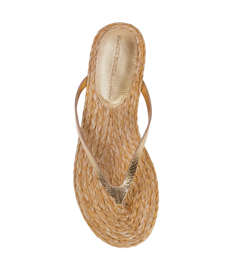 A single woven straw thong sandal with a metallic gold strap, shown from a top-down view - Alaya Sandal in Gold Metallic