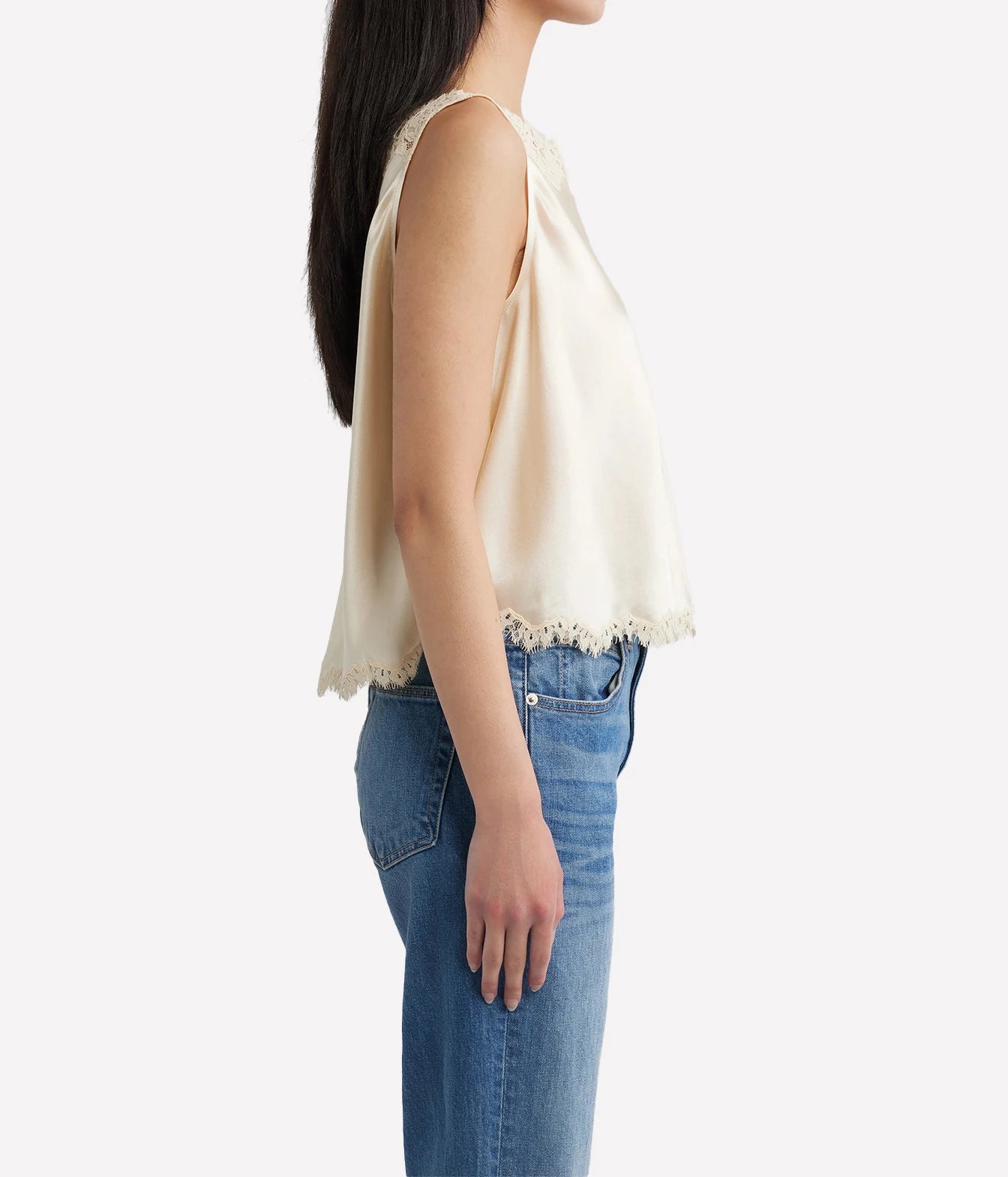 A woman shown from the side wearing a sleeveless cream-colored top with a delicate lace trim and blue jeans, set against a plain light gray background.