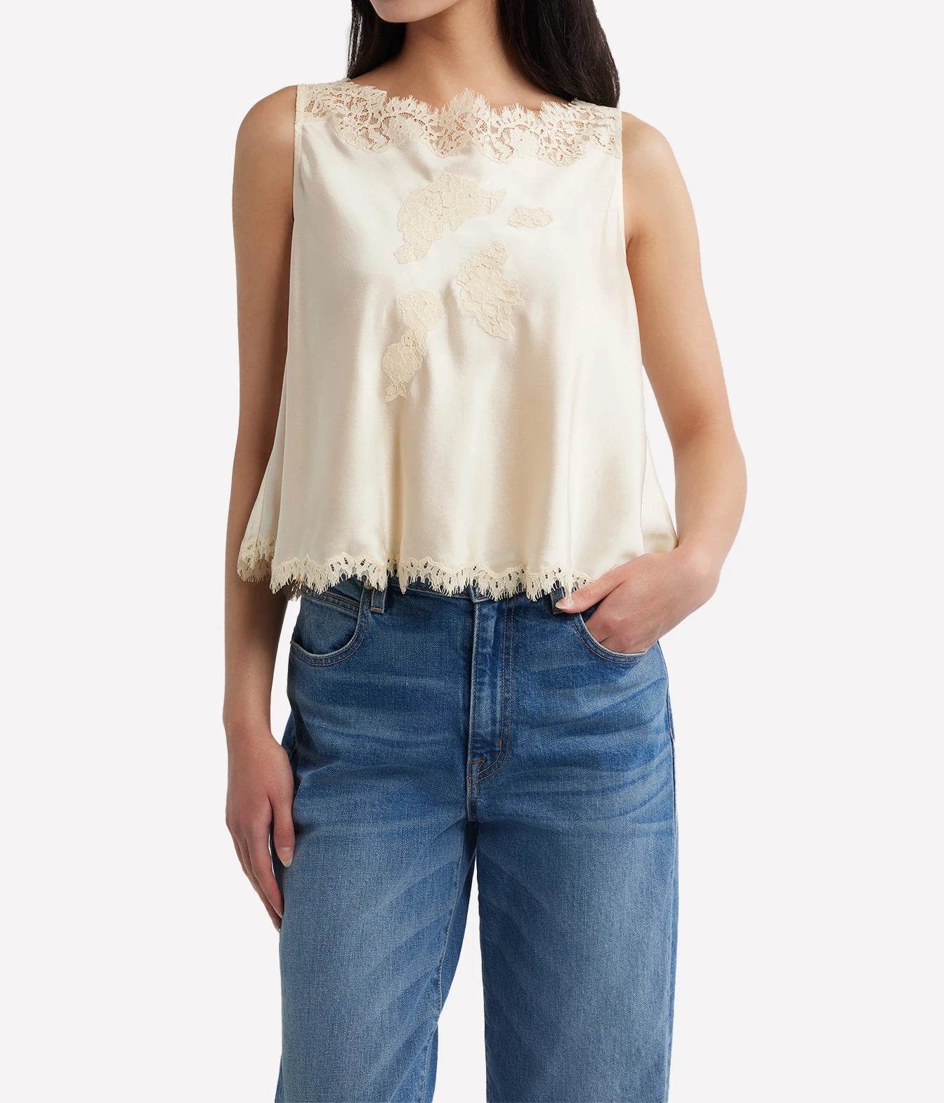 A woman wearing a sleeveless cream satin top with floral lace embroidery and a scalloped hem, paired with blue jeans, against a plain light gray background.