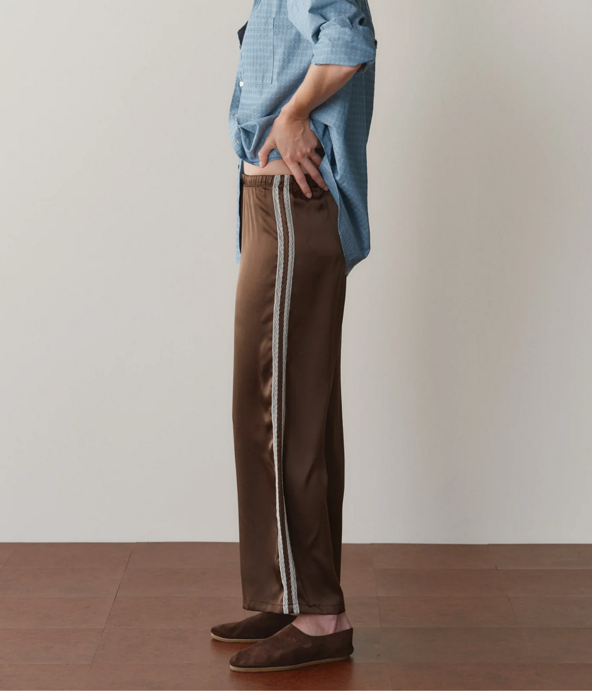 Side profile of a model wearing the DONNI. The Silk Lace Stripe Crop Pant in Espresso, showing the double lace side stripe and straight cropped leg, with a blue printed shirt and brown suede flats