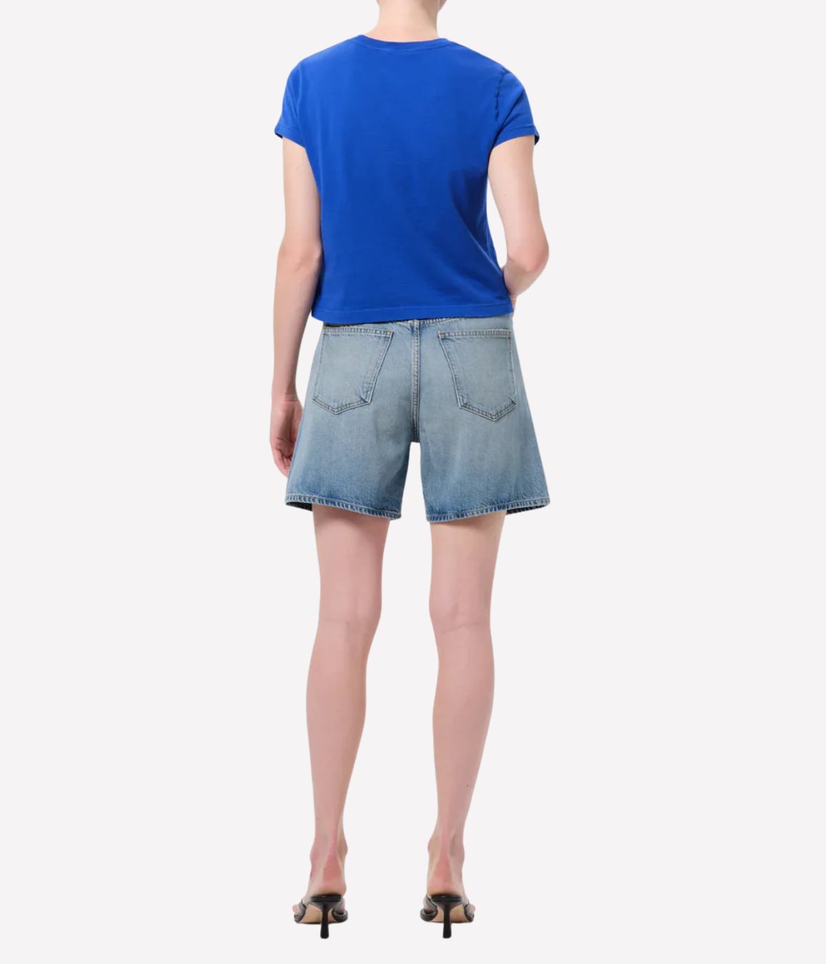 A model shown from the back wearing a bright blue short-sleeve t-shirt and light-wash denim shorts, Agolde Dame Short in Reset