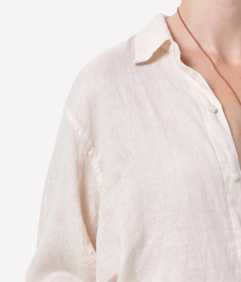 Close-up of a person wearing a light cream linen button-up shirt, showing the collar, textured fabric, and a thin red cord necklace against a plain white background.
