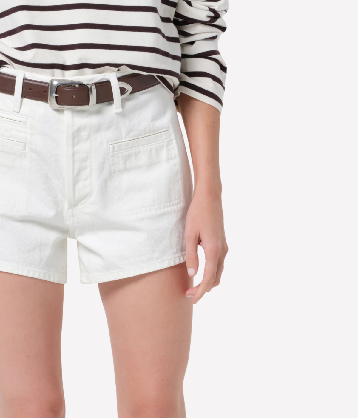 A close-up of a person wearing white denim shorts with a brown leather belt, a black-and-white striped long-sleeve top, and light skin against a plain light gray background.