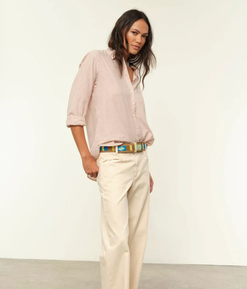 A woman modeling a light pink linen button-down shirt with rolled sleeves, paired with cream-colored trousers and a colorful patterned belt, standing against a plain white studio background.