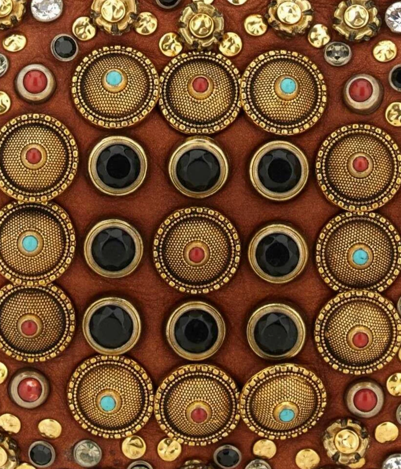 A close-up of a brown leather or suede surface covered with decorative round and oval studs and buttons in gold-tone metal, some with turquoise and red stones and others with black faceted stones.