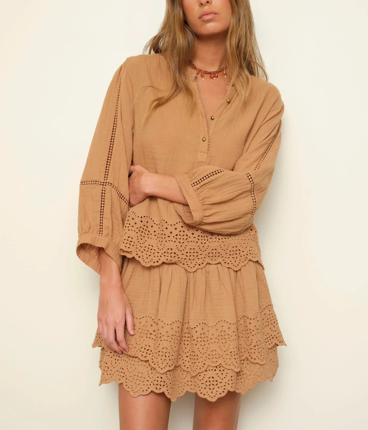 A woman wearing a tan bohemian-style eyelet mini dress with tiered ruffles and long bell sleeves, photographed against a plain off-white background.