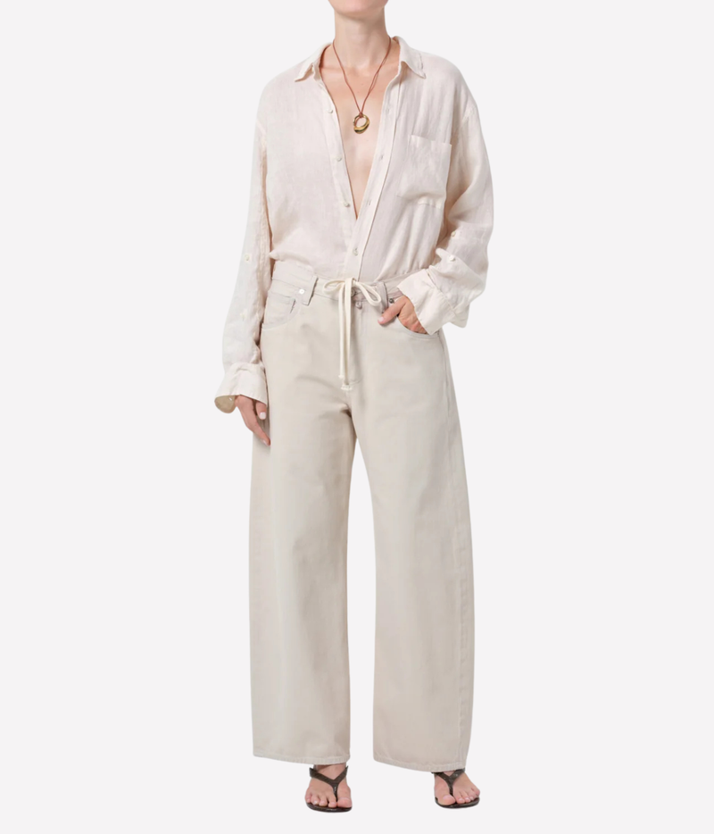 A model wearing a light beige linen two-piece set consisting of a long-sleeve button-down shirt and matching wide-leg trousers, styled with a gold pendant necklace and black sandals against a plain white background.