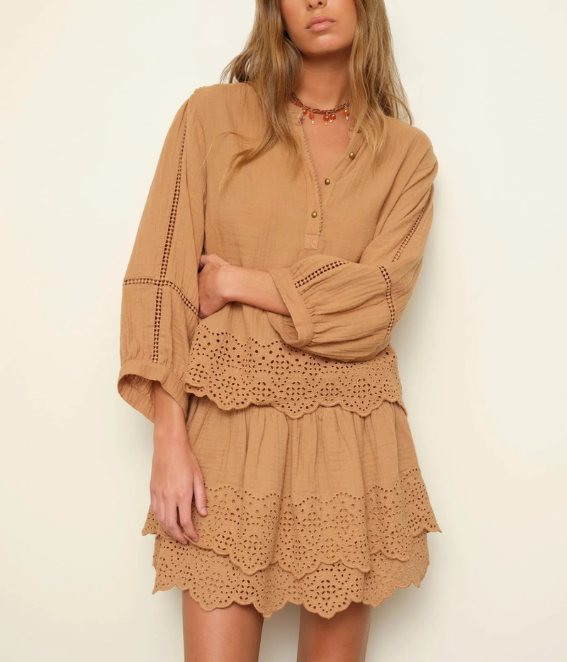 A woman wearing a tan bohemian-style eyelet mini dress with tiered ruffles and long bell sleeves, photographed against a plain off-white background.