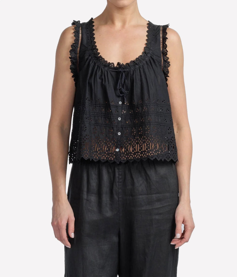 A woman wearing a black sleeveless eyelet lace top with ruffled trim and a drawstring neckline, paired with black pants, against a light gray studio background.