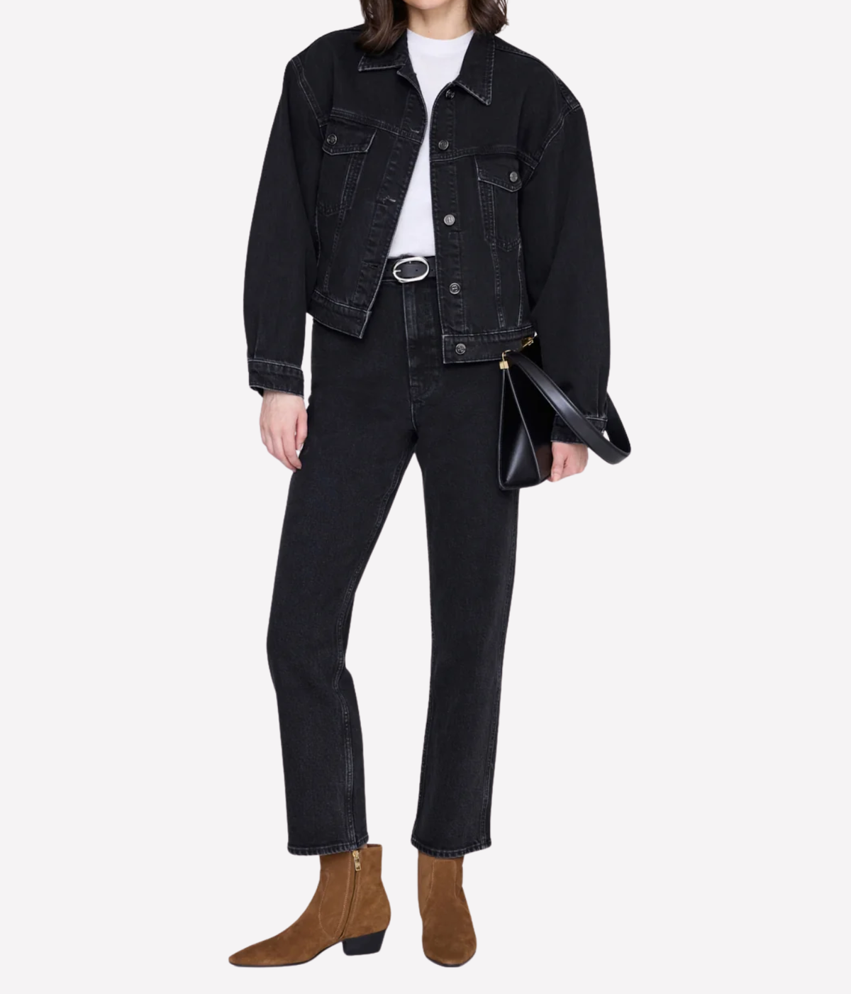 A model is shown from the waist down wearing a black denim jacket, black jeans, a white t-shirt, brown suede ankle boots, and holding a black leather handbag against a light gray background.