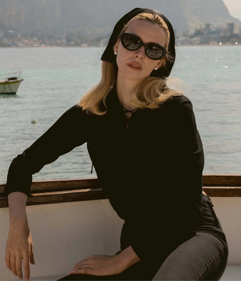 A woman wearing a black long-sleeve top, black pants, a black headscarf, and black sunglasses
