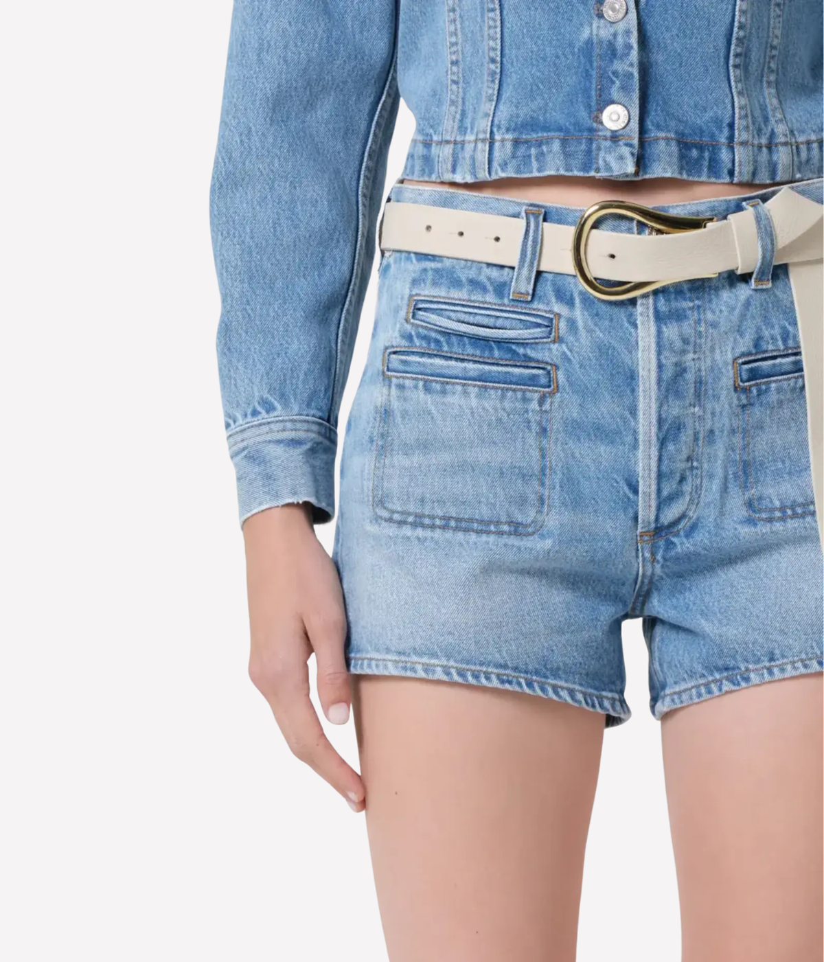 Close-up of a model wearing light-wash blue denim shorts with front patch pockets, a cream belt with a gold buckle, and a matching cropped denim jacket against a plain light gray background.