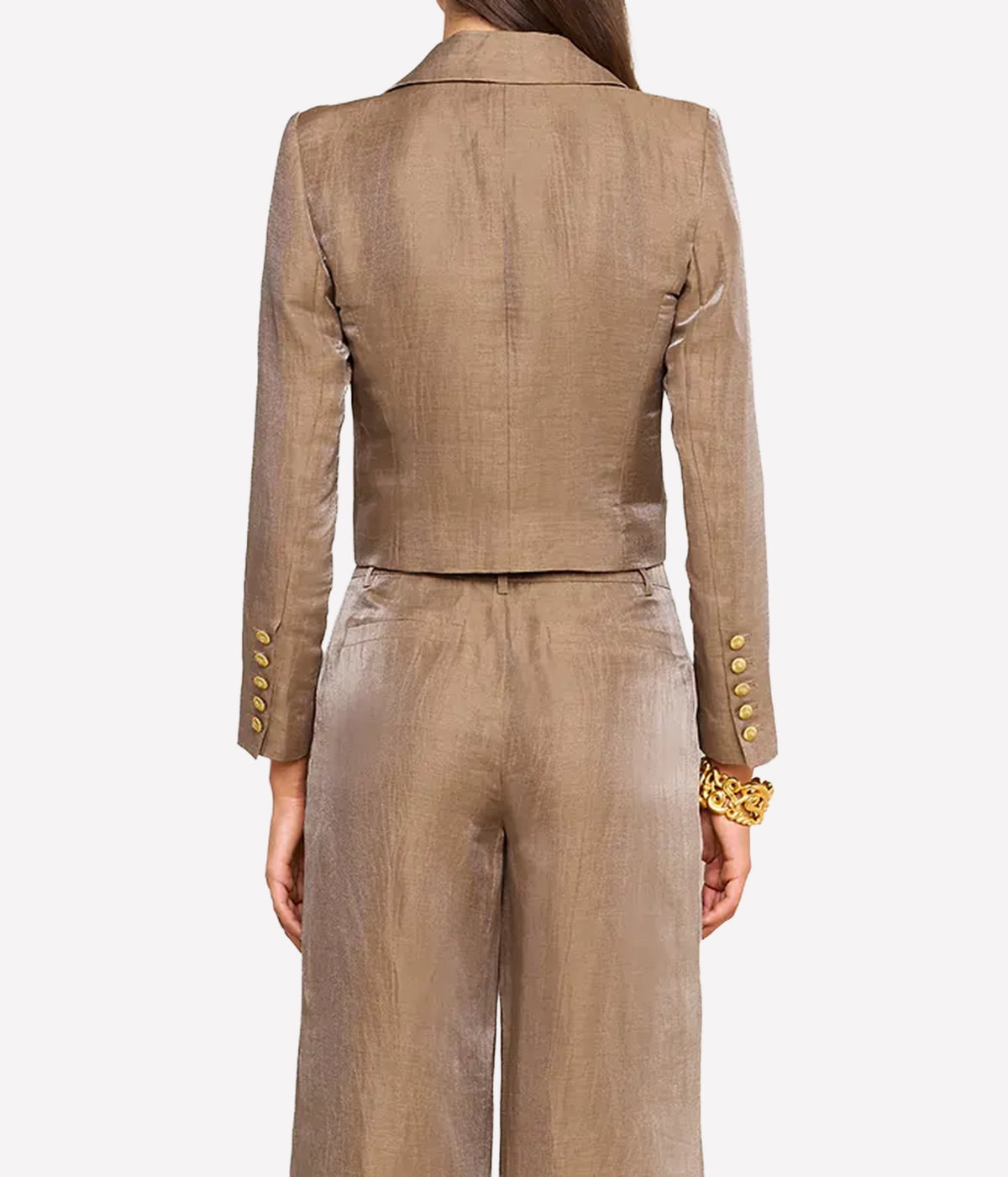 A model shown from the back wearing a matching cropped blazer and wide-leg trousers set in a shimmering taupe-beige fabric, featuring gold buttons on the sleeves and a gold patterned cuff bracelet.