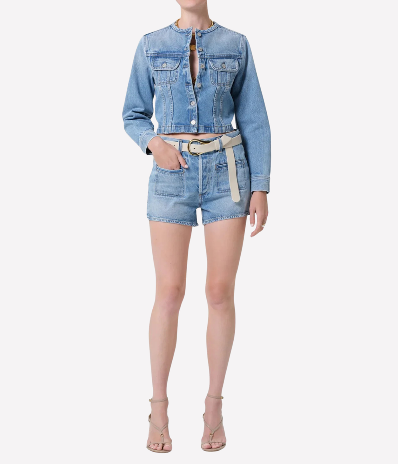 A model wearing a light blue denim two-piece set consisting of a cropped long-sleeve button-front jacket with chest pockets and matching denim shorts, styled with a beige belt and nude high-heeled sandals against a light gray background.
