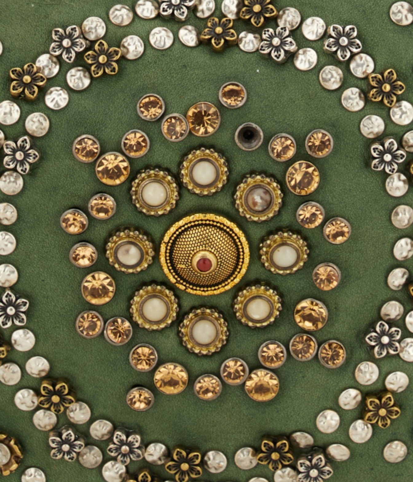 A flat-lay arrangement of decorative craft embellishments on a textured green background, featuring a central gold-toned medallion surrounded by concentric rings of gold, silver, and clear rhinestones and floral-shaped beads.