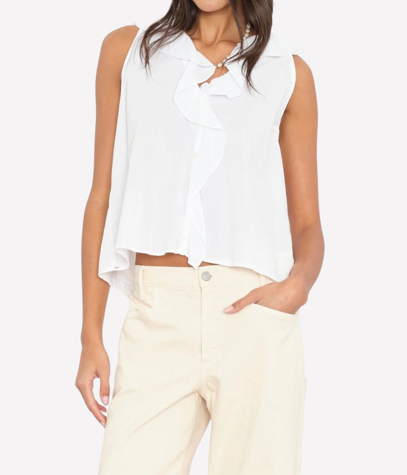 A woman wearing a sleeveless white blouse with a ruffled collar and front detail, paired with cream-colored pants, against a plain light gray background.