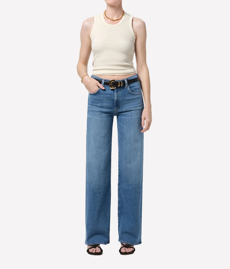 Full-length front view of a model wearing the Citizens Of Humanity Annina 33" Jean in Stella with a cream ribbed tank top tucked in, a black leather belt, a gold necklace and dark strappy sandals