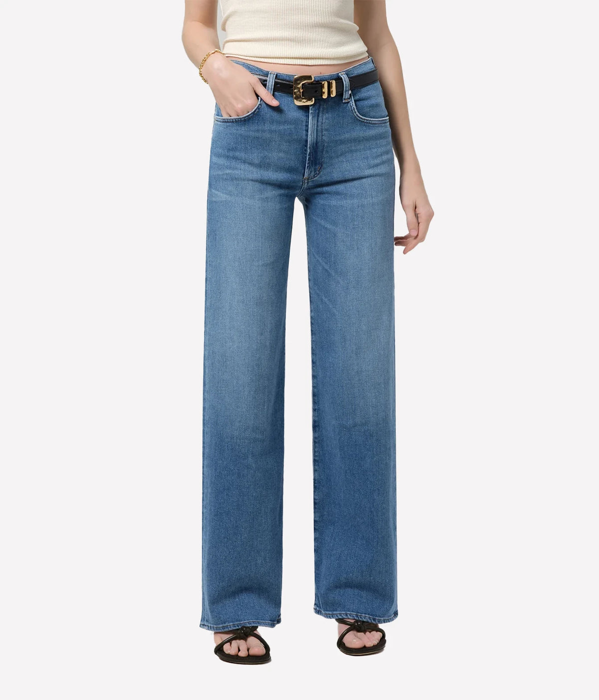 Front view of a model wearing the Citizens Of Humanity Annina 33" Jean in Stella with a cream tank top tucked in and a black leather belt with a gold buckle, one hand in the front pocket, showing the high waist and full-length wide leg, paired with dark sandals