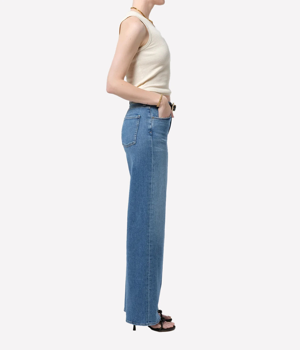 Side profile of a model wearing the Citizens Of Humanity Annina 33" Jean in Stella with a cream tank top and one hand in the pocket, showing the straight wide leg falling to the floor over dark heeled sandals