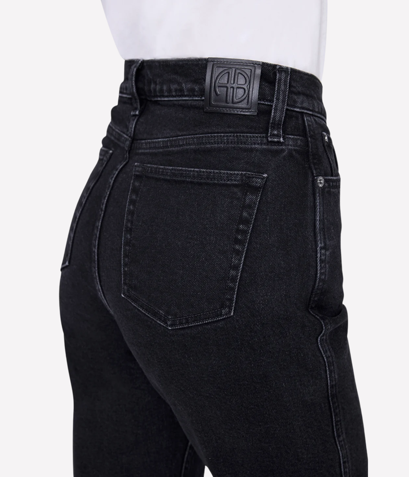 Close-up rear view of a person wearing black denim jeans with a white shirt at the waist, showing the back pocket, stitching, and a black leather brand patch on the waistband.