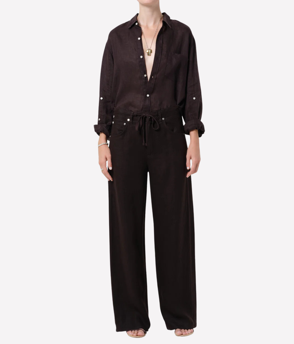 A model wearing a dark brown wide-leg jumpsuit with a matching long-sleeve button-up shirt, styled with a gold pendant necklace and sandals against a plain white background.
