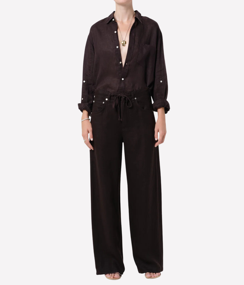 A model wearing a dark brown wide-leg jumpsuit with a matching long-sleeve button-up shirt, styled with a gold pendant necklace and sandals against a plain white background.