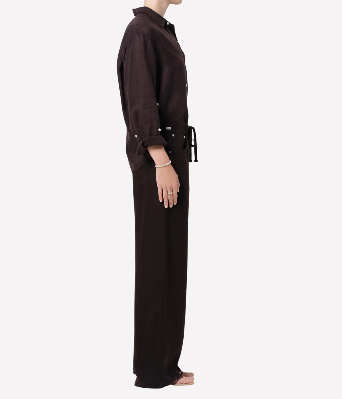 A model shown in profile wearing a dark brown long-sleeve button-up shirt and matching wide-leg trousers with a tie belt, against a plain light gray background.