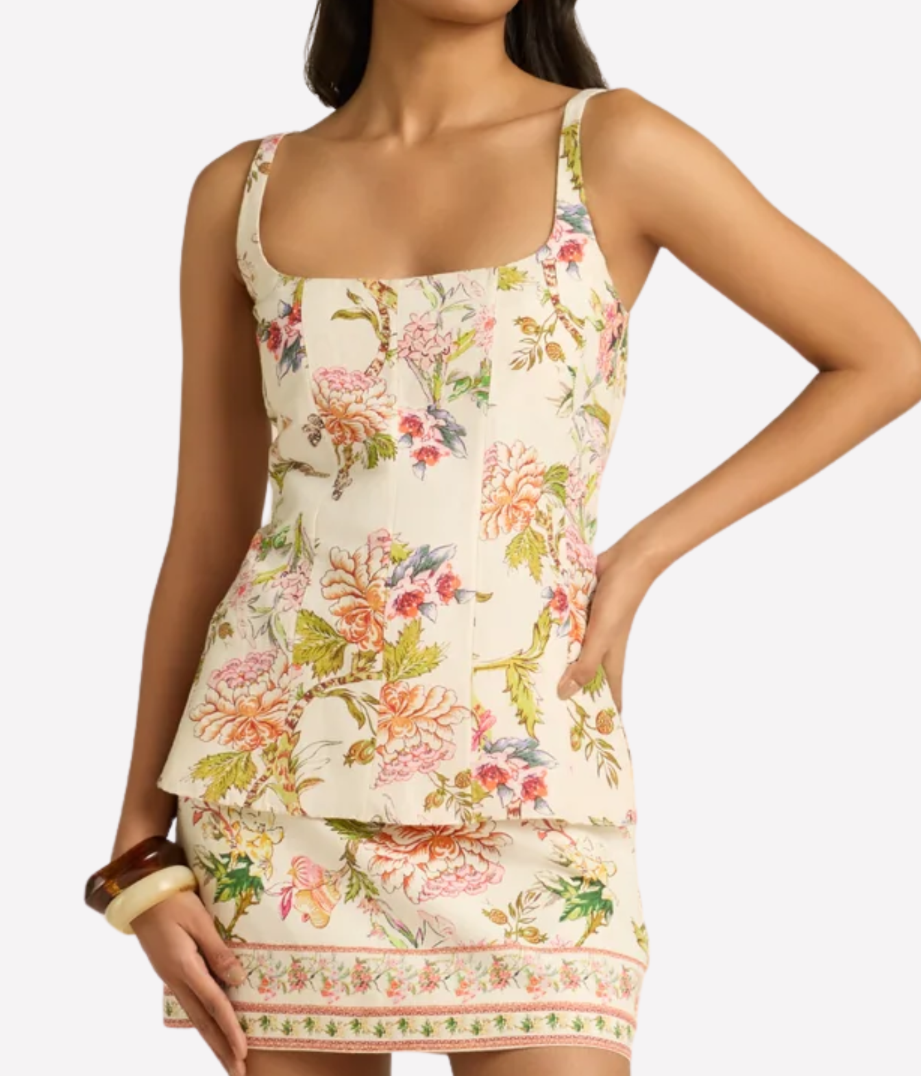 A woman wearing a sleeveless peplum-style mini dress with a floral toile print on a cream background, featuring pink, green, and orange flowers. The image is cropped from the neck down against a light gray studio background.