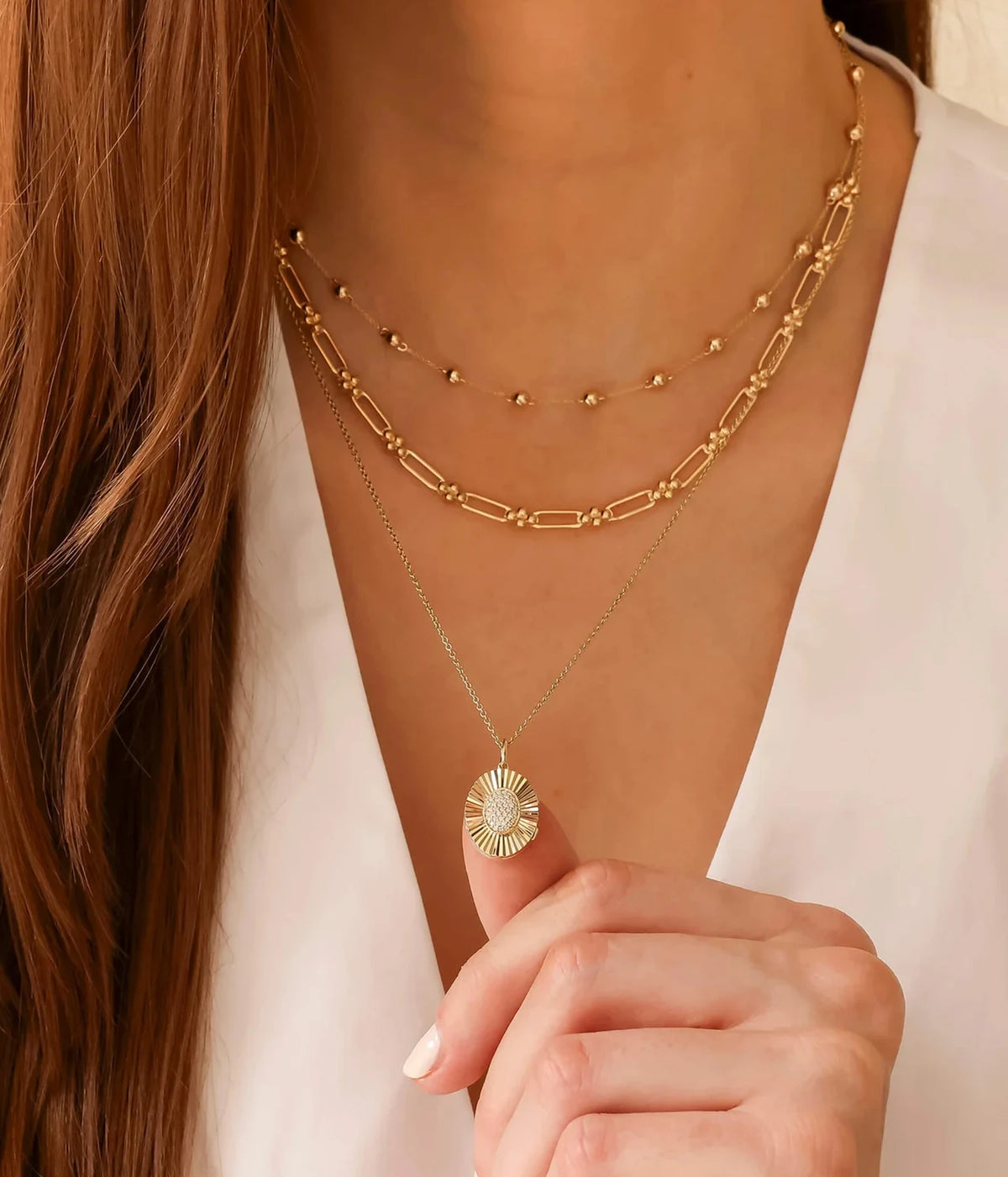Model in an open-collar white shirt holding the Dana Rebecca Designs Teddi Paige Oval Locket Necklace in 14k Yellow Gold, layered with gold link chains