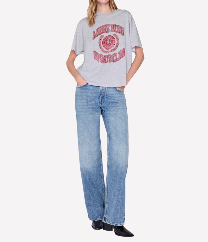 A model wearing a light gray oversized t-shirt with a distressed red graphic and light-wash blue jeans, standing against a plain light gray background.