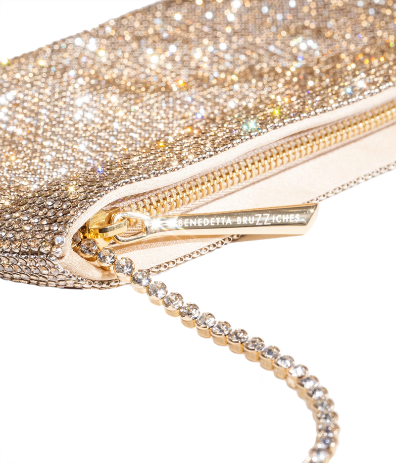 Close-up of a glittery gold evening clutch bag with a rhinestone-embellished chain strap and a gold zipper pull engraved with the brand name Benedetta Bruzziches.