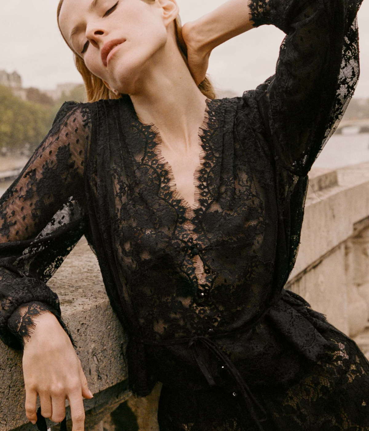 Close-up of a model wearing the DÔEN Amaline Top In Black with one arm raised, showing the plunging eyelash-lace V-neckline, sheer lace sleeves and tied waist