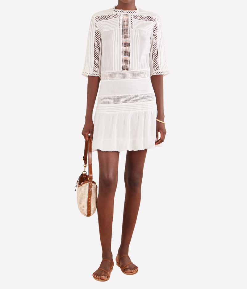A model wearing the Isabel Marant Paige Dress In White, a crochet elbow-length sleevemini dress with brown leather sandals and carrying a woven straw handbag