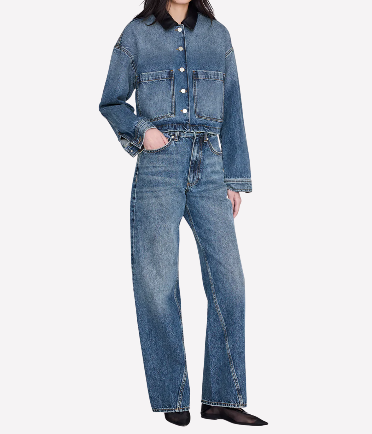 A model wearing a cropped blue denim jacket with a contrasting black collar and matching wide-leg blue denim jeans, styled with black pointed shoes against a light gray background.