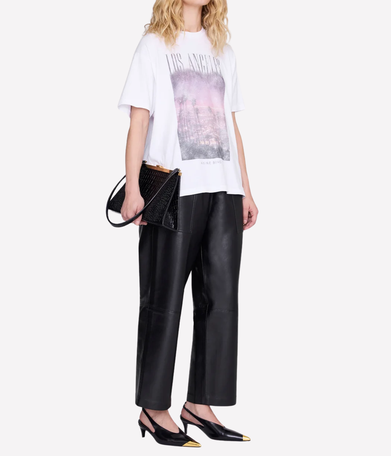 Model wearing the Anine Bing Cade Tee Los Angeles In White untucked over black leather straight-leg trousers, holding a black woven leather shoulder bag and wearing black slingback heels with gold toe caps
