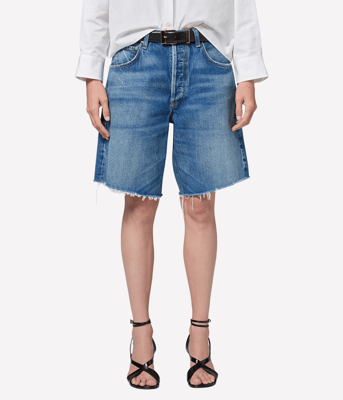 Close-up front view of a model wearing the Citizens of Humanity Ayla Short in Chaya with a black leather belt and a tucked white shirt, showing the faded medium wash, button fly, knee length and frayed raw hems, with black strappy heeled sandals