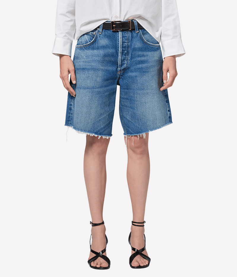 Close-up front view of a model wearing the Citizens of Humanity Ayla Short in Chaya with a black leather belt and a tucked white shirt, showing the faded medium wash, button fly, knee length and frayed raw hems, with black strappy heeled sandals