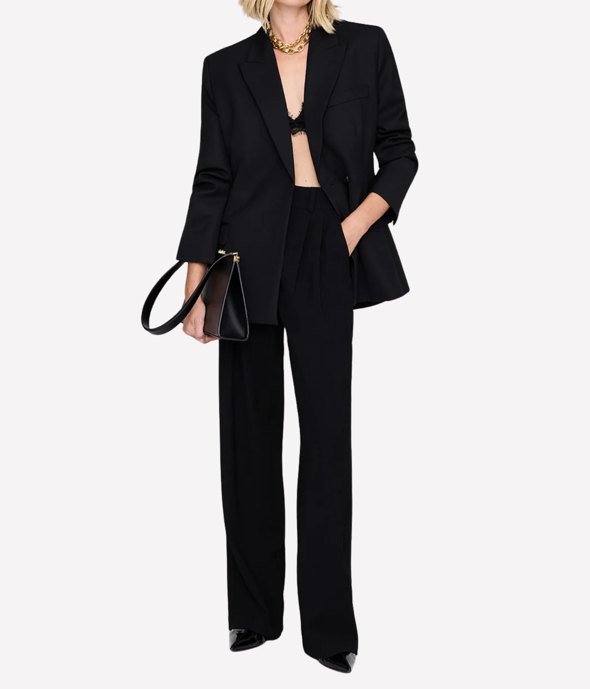 Model wearing the Anine Bing Aretha Blazer in Black open over a black bralette with matching wide-leg trousers, gold chain necklace and a dark leather handbag