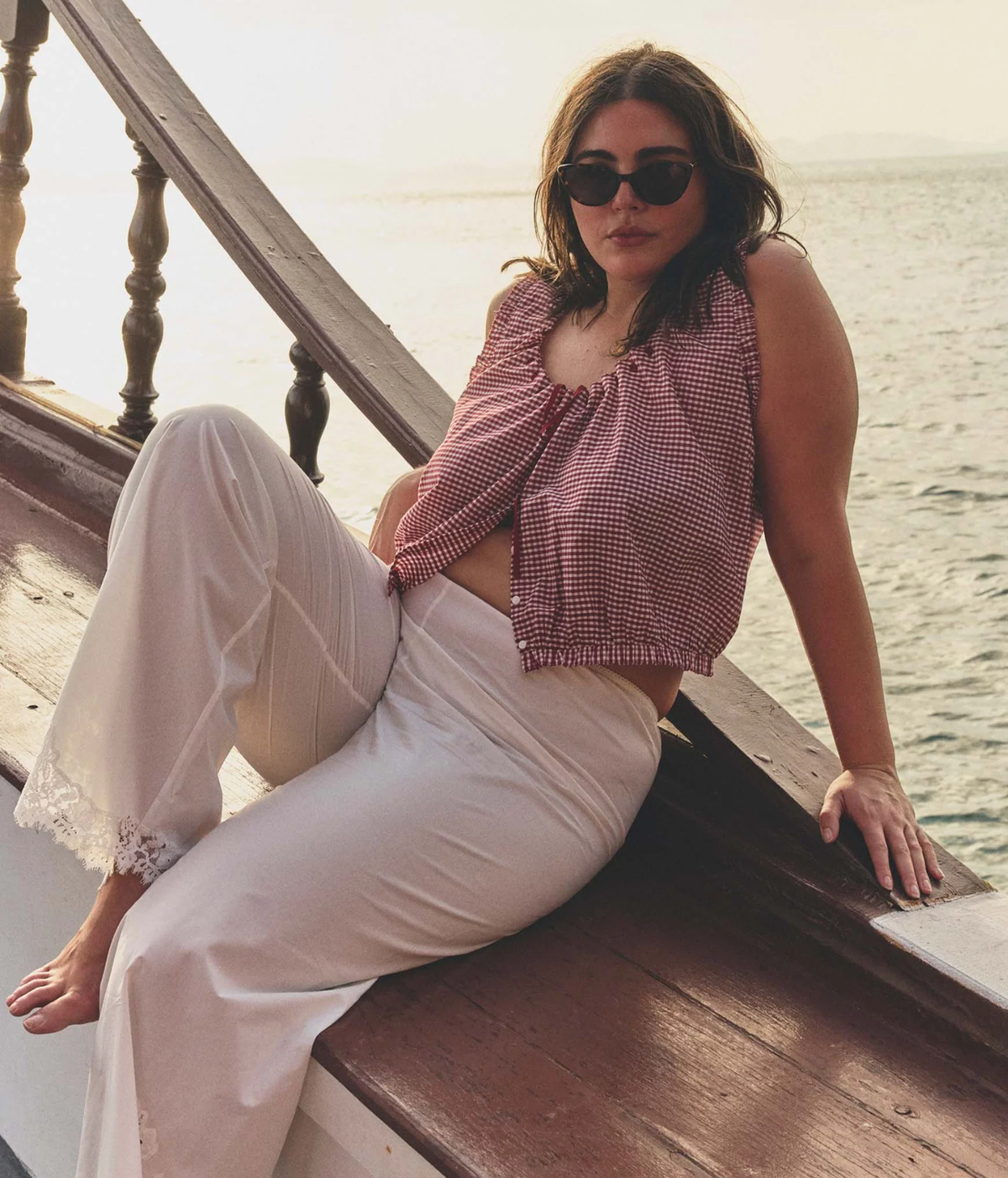 Model wearing the DOEN Traveler Top in Mulberry La Maddalena Gingham with white trousers by the water.