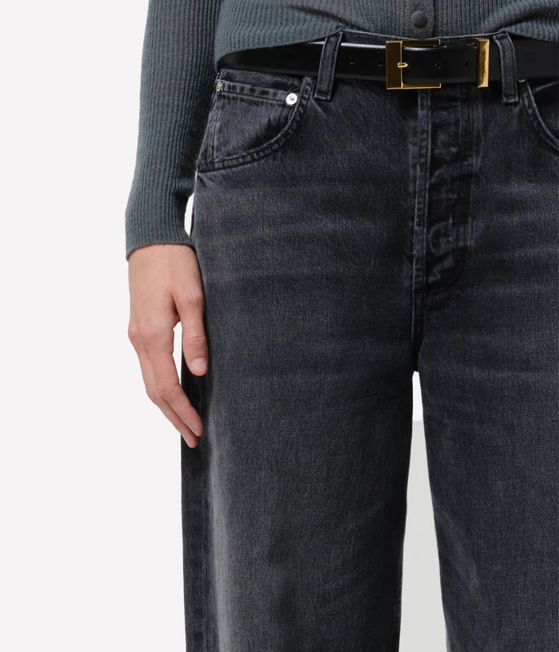 Close-up of a person wearing dark gray washed denim jeans with a black leather belt featuring a gold-tone rectangular buckle, paired with a ribbed dark gray long-sleeve top against a plain light background.