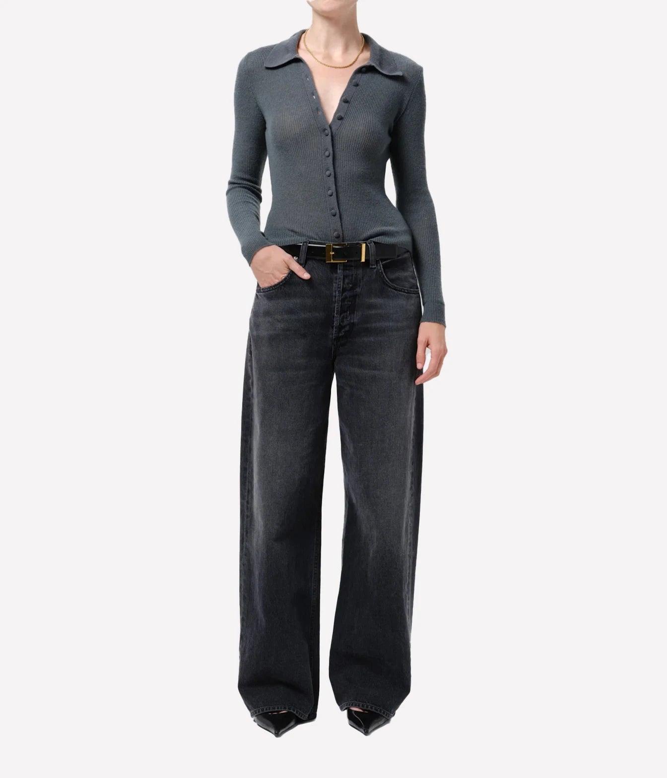 A model wearing a dark gray long-sleeve button-up knit top and wide-leg dark gray jeans with a gold-tone belt, photographed against a plain light gray studio background.