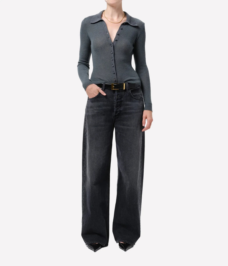 A model wearing a dark gray long-sleeve button-up knit top and wide-leg dark gray jeans with a gold-tone belt, photographed against a plain light gray studio background.