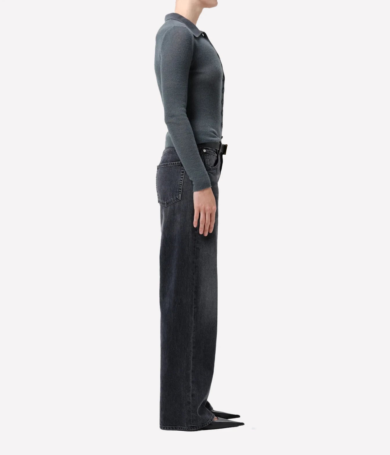 A side profile view of a model wearing a dark charcoal gray long-sleeve knit polo shirt tucked into dark gray wide-leg jeans, paired with black pointed-toe flats, against a solid off-white background.