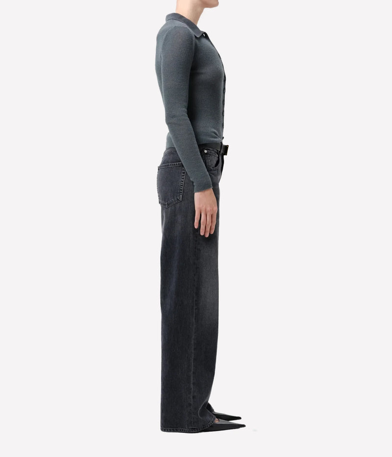 A side profile view of a model wearing a dark charcoal gray long-sleeve knit polo shirt tucked into dark gray wide-leg jeans, paired with black pointed-toe flats, against a solid off-white background.