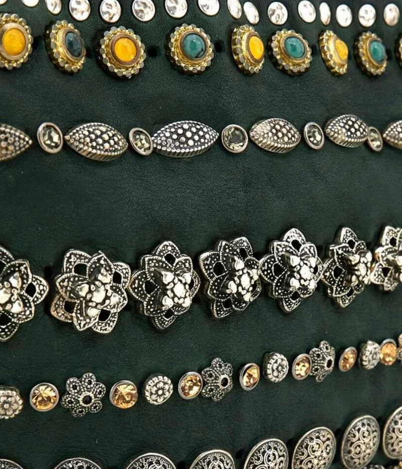 A close-up of a dark green fabric surface decorated with multiple horizontal rows of ornate metal studs, brooches, and embellished buttons in silver, gold, and turquoise tones.