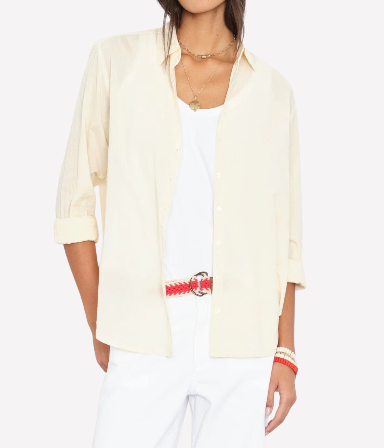 A woman wearing a light cream-colored button-down shirt over a white tank top and white pants, accessorized with a red and white braided belt and gold necklaces, against a plain white background.