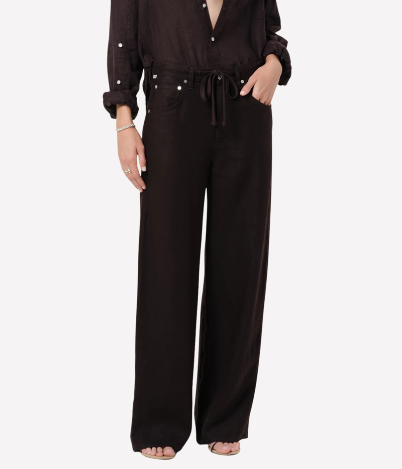 A model wearing a dark brown wide-leg pants jumpsuit with a drawstring waist and decorative silver buttons, shown from the chest down on a light gray background.